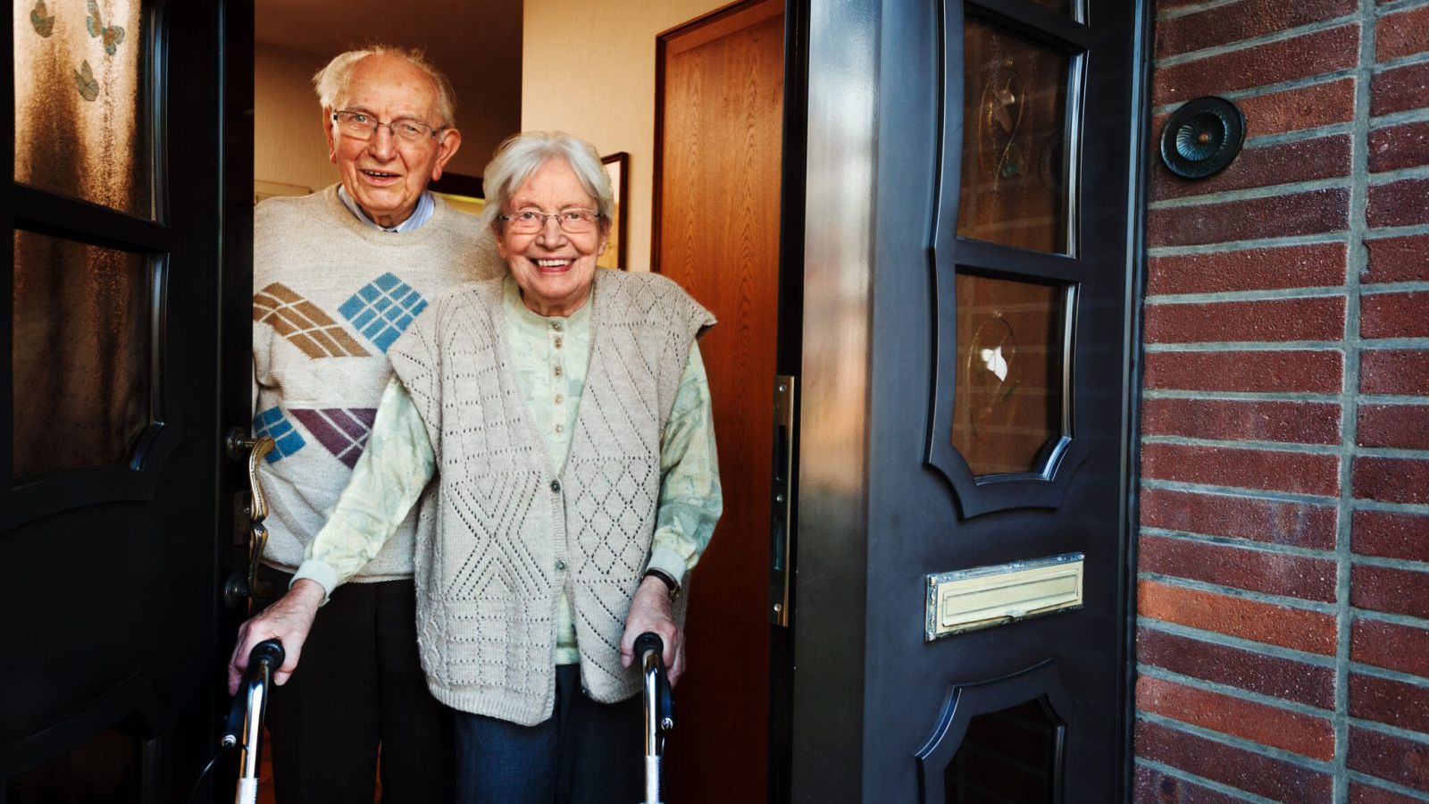 Smiling elderly couple standing at the front door of a residential board and care home, with the woman using a walker and the man standing behind her, representing a safe, welcoming, and supportive senior living environment.