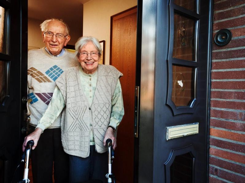 Smiling elderly couple standing at the front door of a residential board and care home, with the woman using a walker and the man standing behind her, representing a safe, welcoming, and supportive senior living environment.