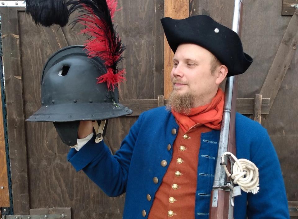 Person i historisk dräkt håller en hjälm och ett gevär.