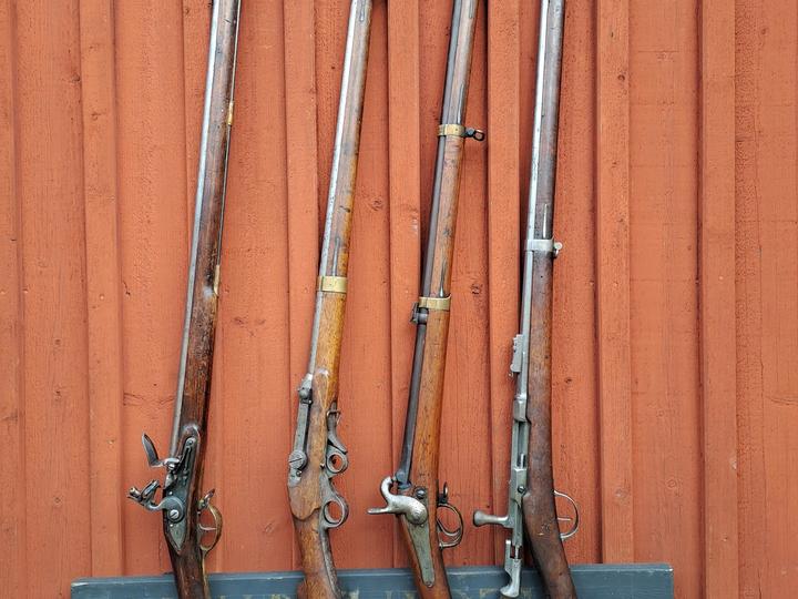 Fyra historiska långvapen uppställda mot en röd trävägg.