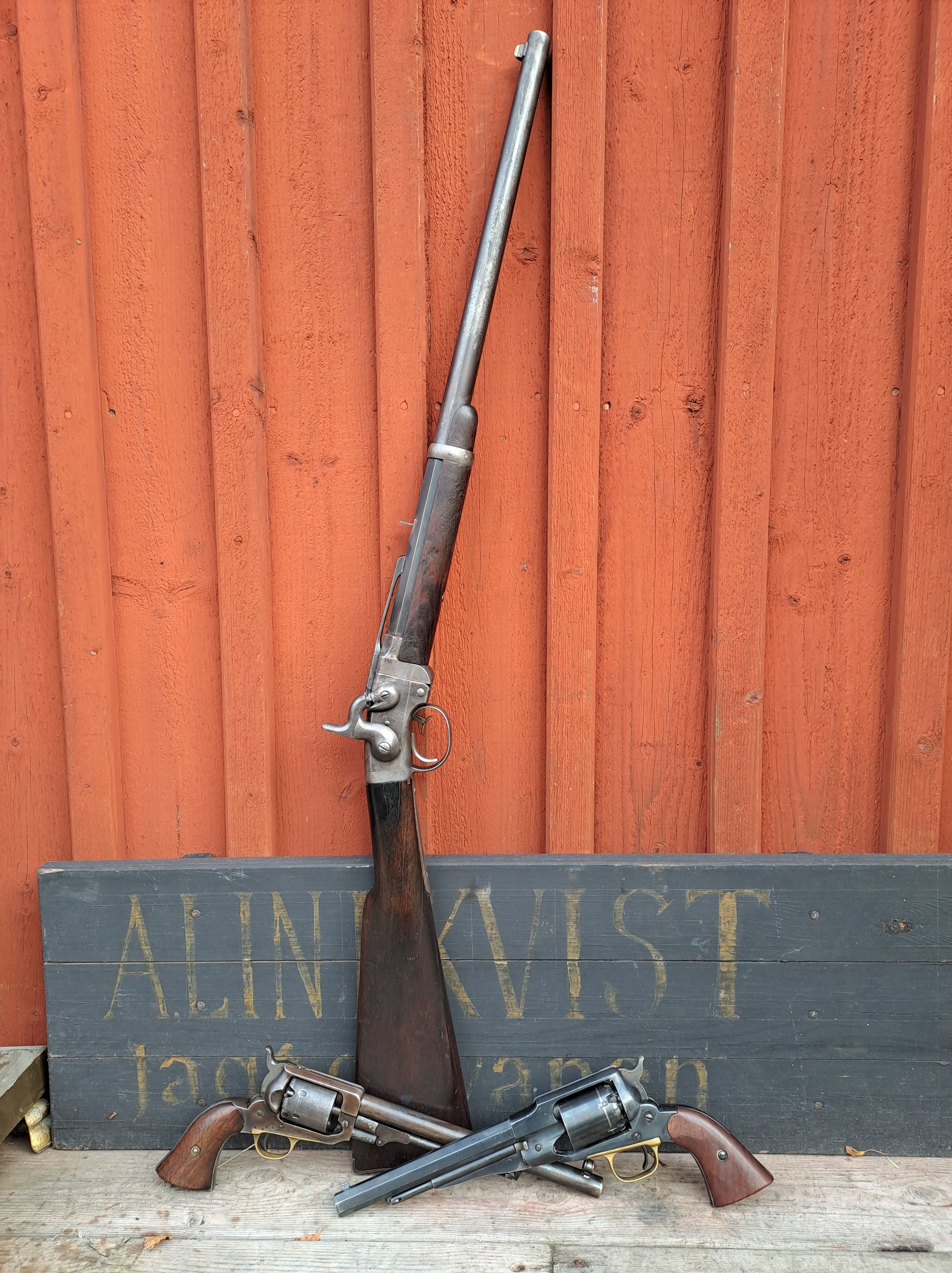 Historiskt gevär och revolvrar uppställda framför en röd trävägg.