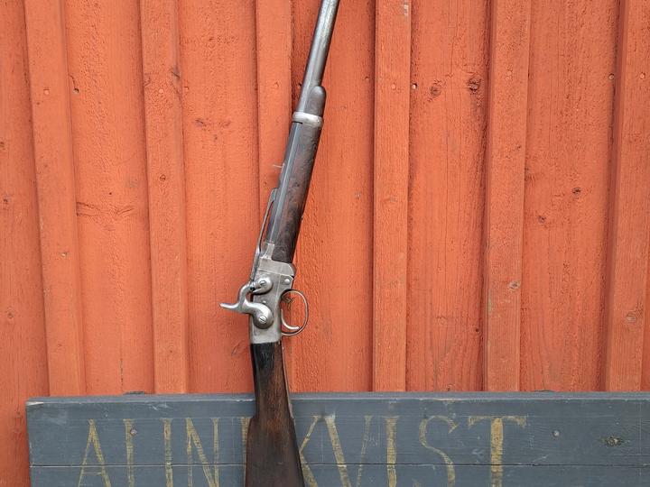 Historiskt gevär och revolvrar uppställda framför en röd trävägg.