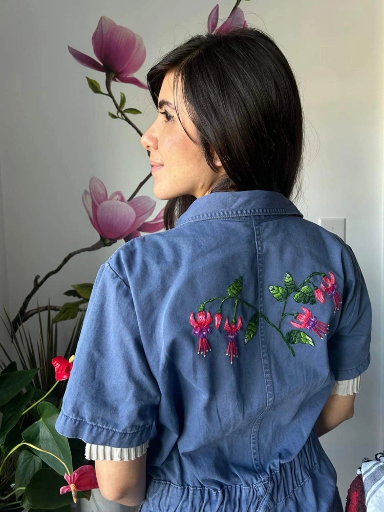 Jess wearing a jean romper with an embroidery of pink fuschias and green leaves in the back.