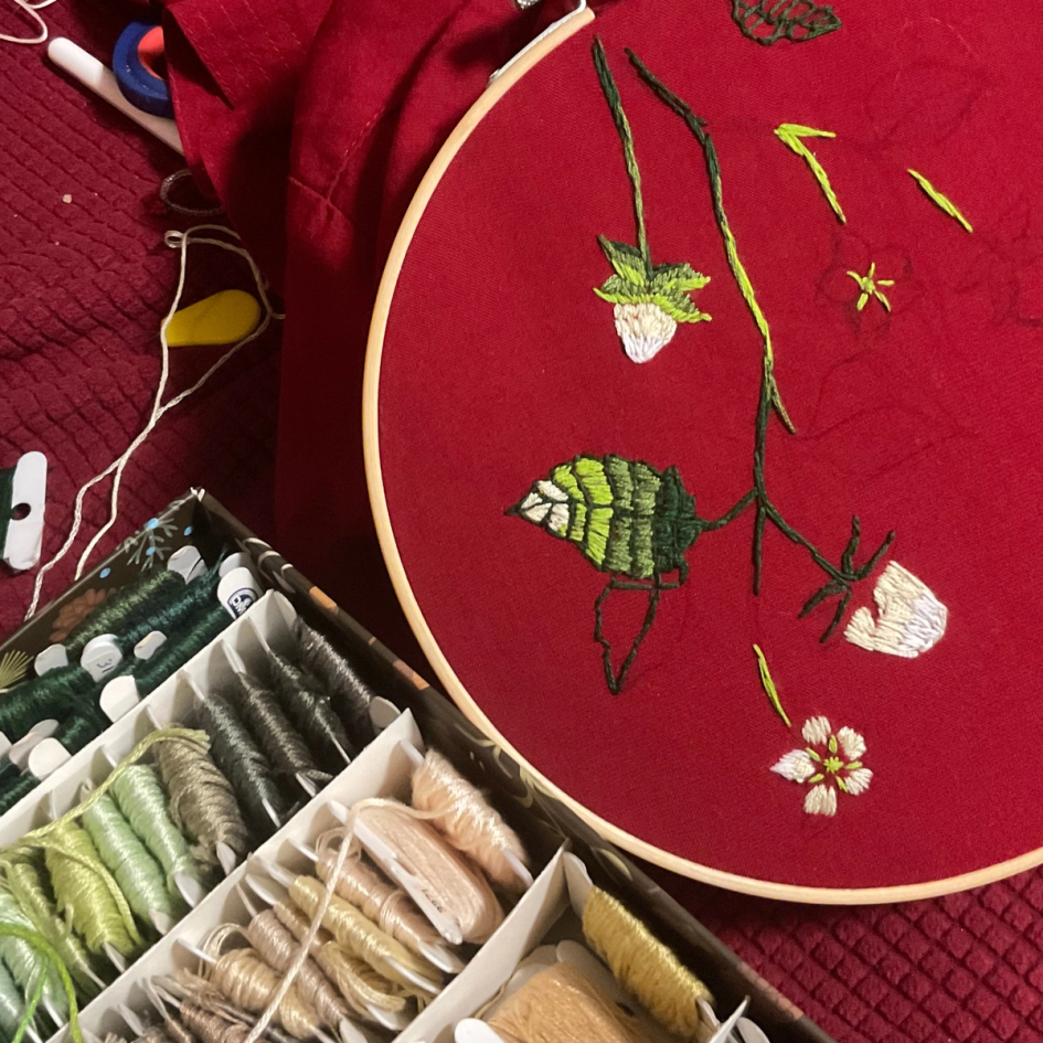 Close up of the process of embroidering pine berries into a red shirt. In the corner there is a box of green and beige threads.
