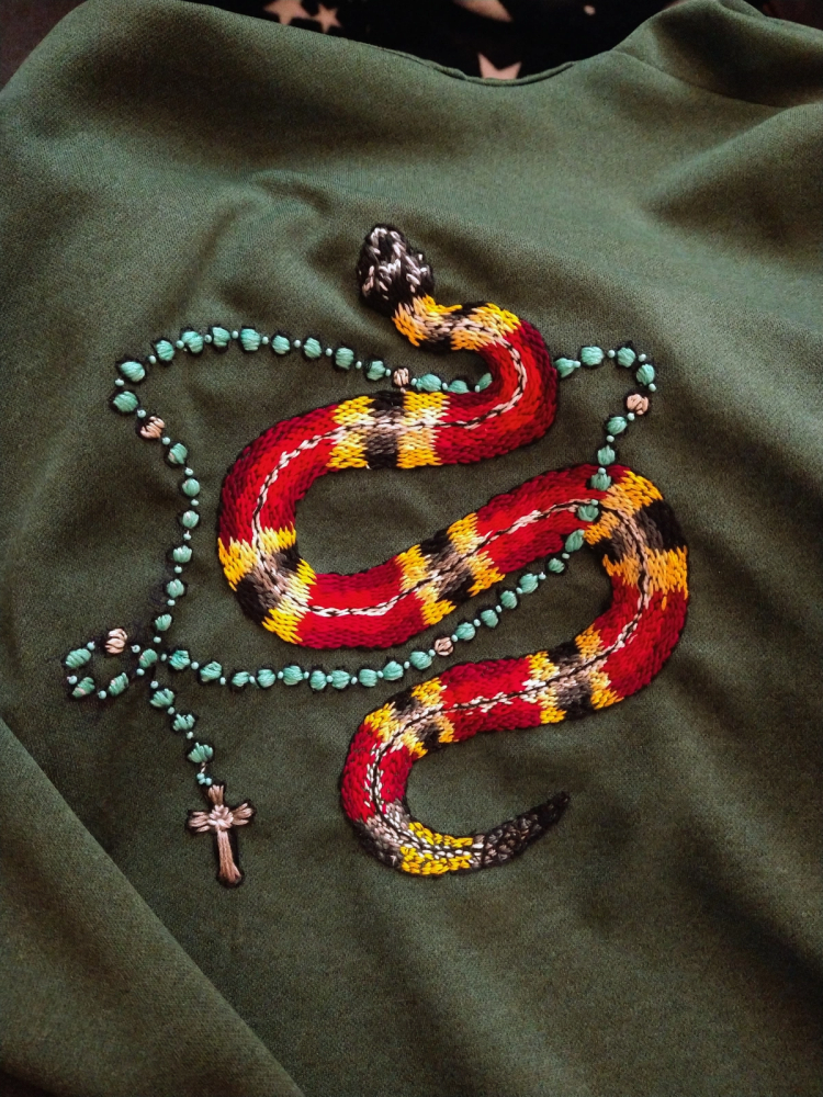 The embroidery of a coral snake wrapped in a rosary on green fabric.