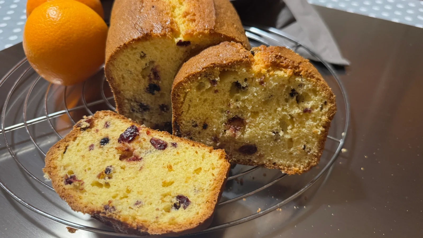 Slices of orange red berry pound cake on a wire rack next to fresh whole oranges on a stainless steel surface.
