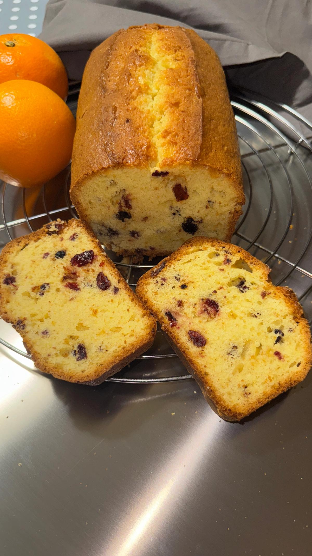 Slices of orange red berry pound cake on a wire rack next to fresh whole oranges on a stainless steel surface.