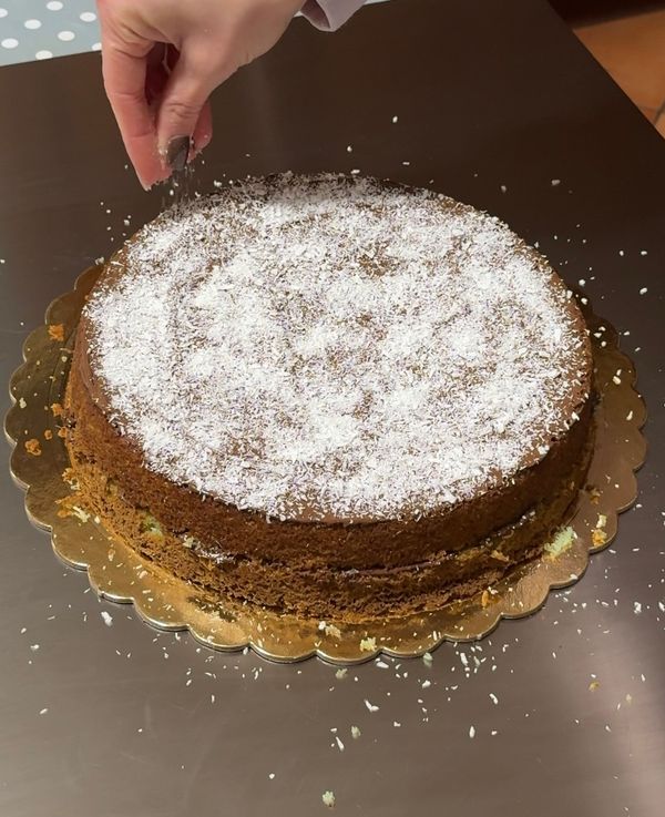 Hand sprinkling desiccated coconut over a coconut Nutella mint cake on a gold board, viewed from above.