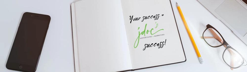 A phone, open book, pencil, and glasses with laptop on table. Book says your success =  jdoc success.