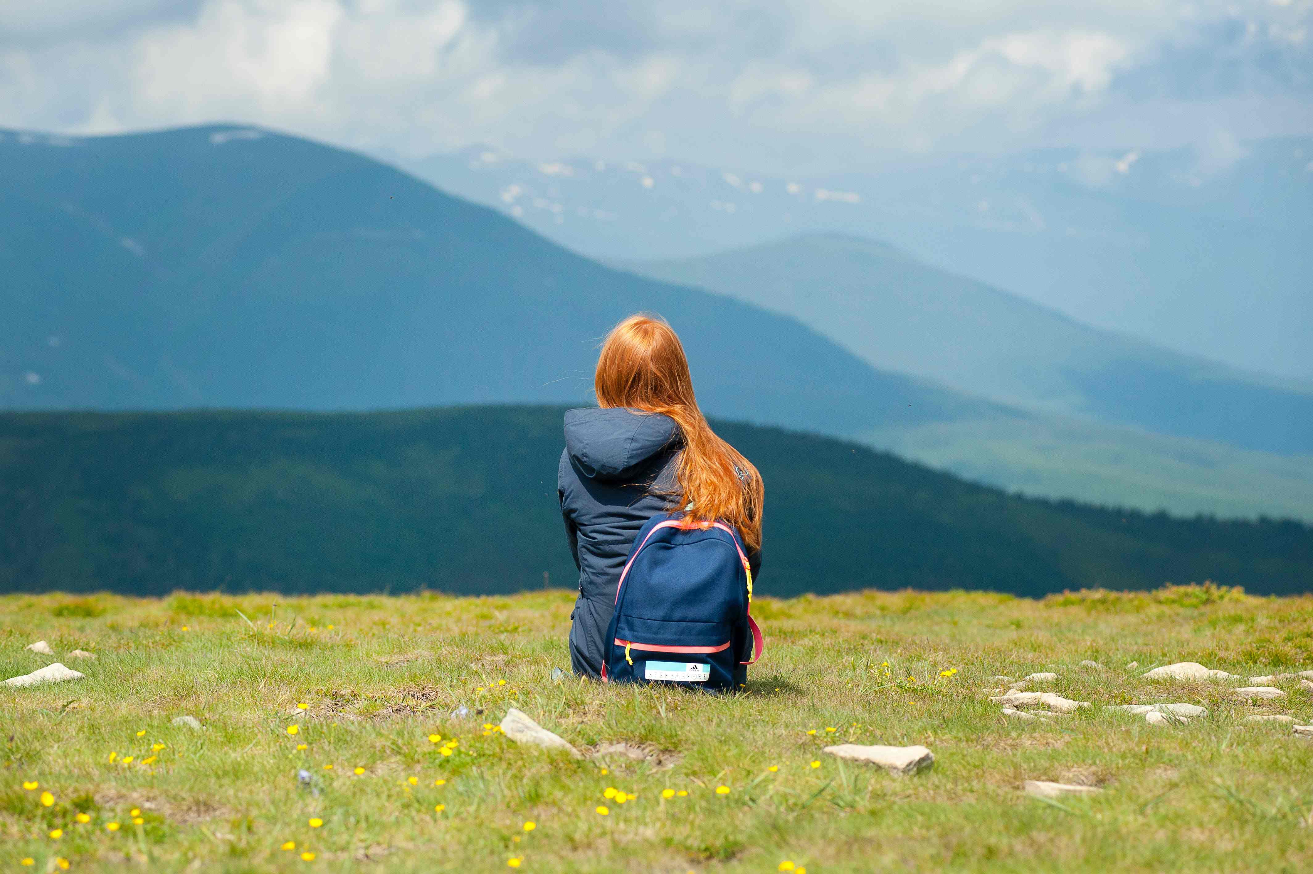 Woman with a backpack sitting in grass, admiring the view of mountains and valleys.