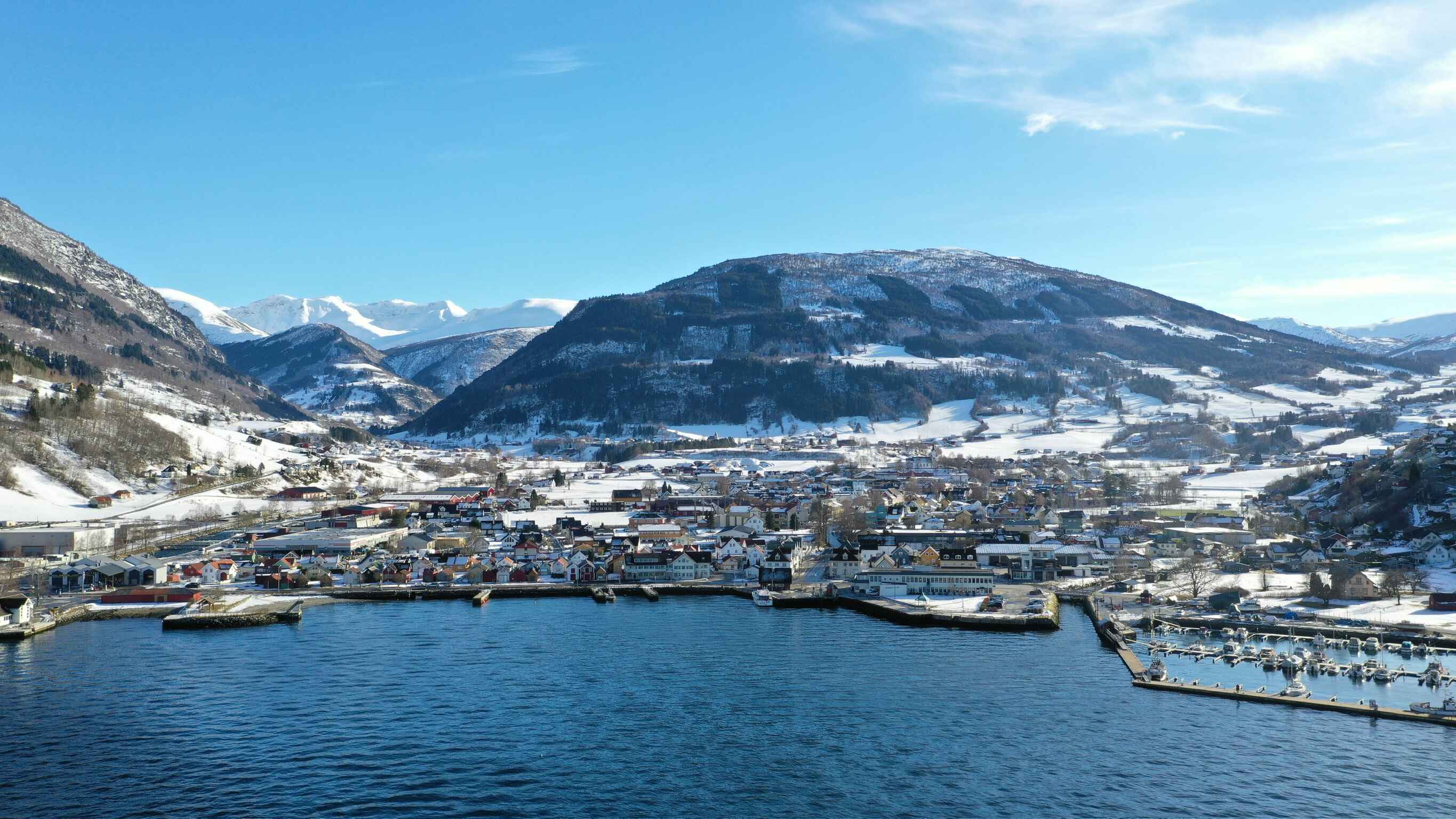 Aerial view of Vikøyri by the fjord with buildings and docks along the water's edge, backed by snow-covered mountains under a clear blue sky.