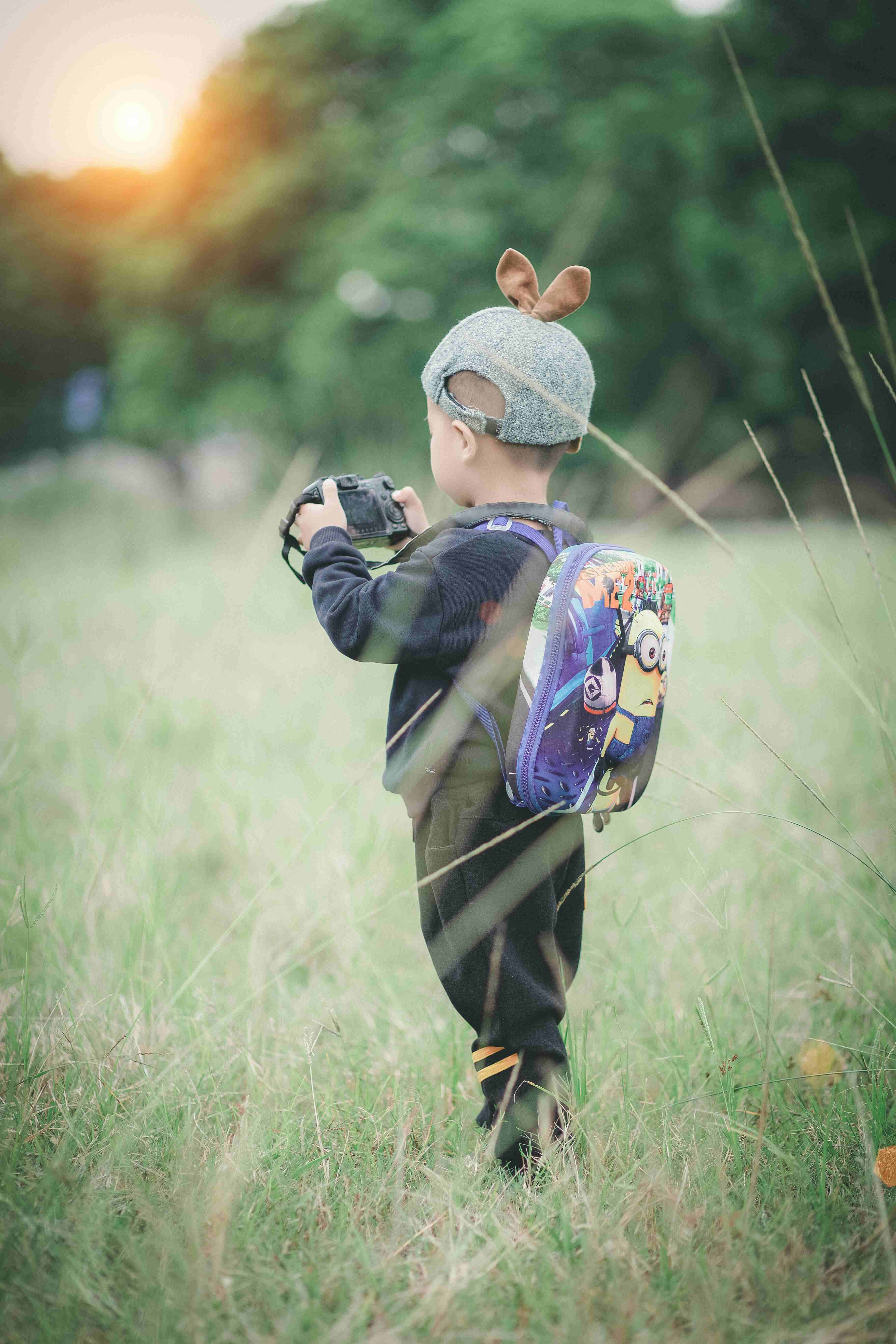 A little boy wearing a backpack and a beanie with two ears on top, holding a camera in a grassy field.