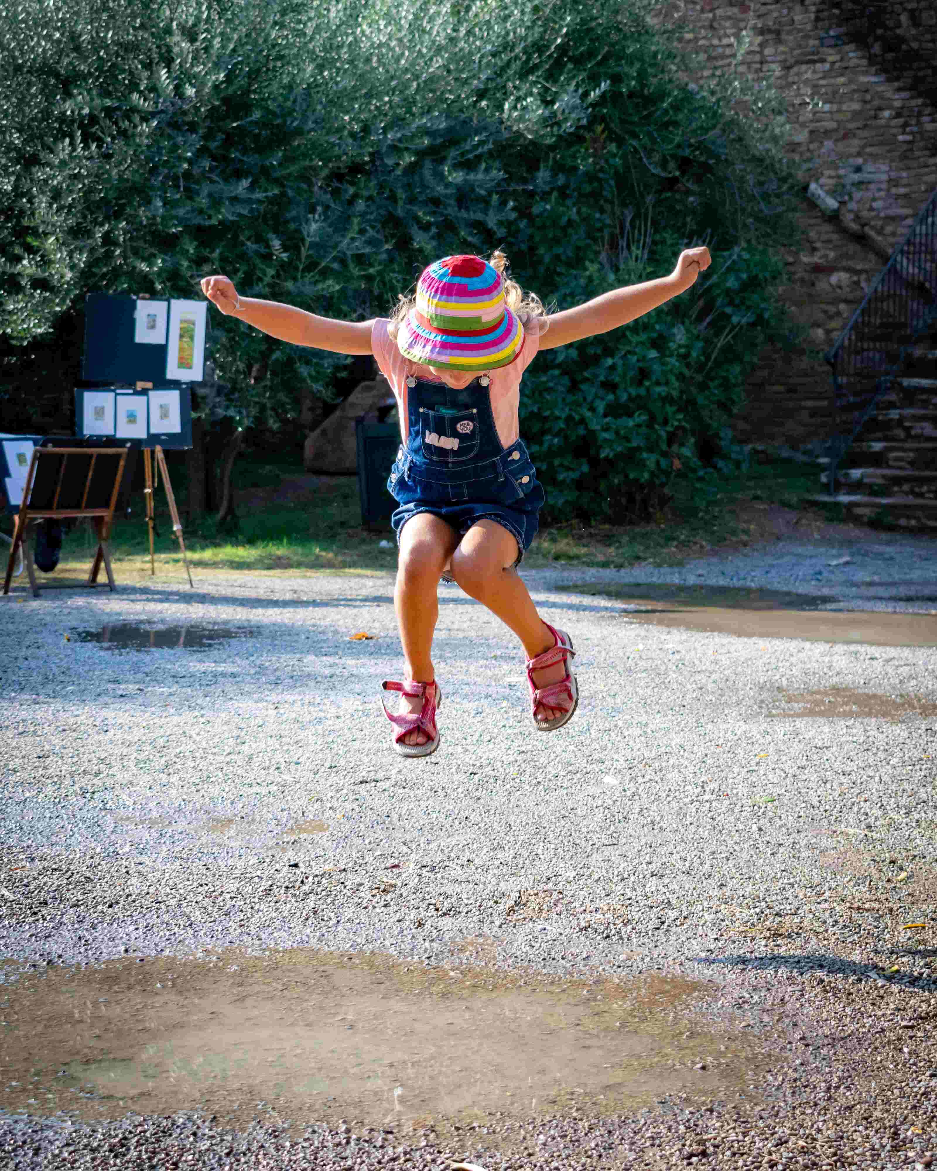 A little girl in a festive hat and jumping joyfully on a dirt path.
