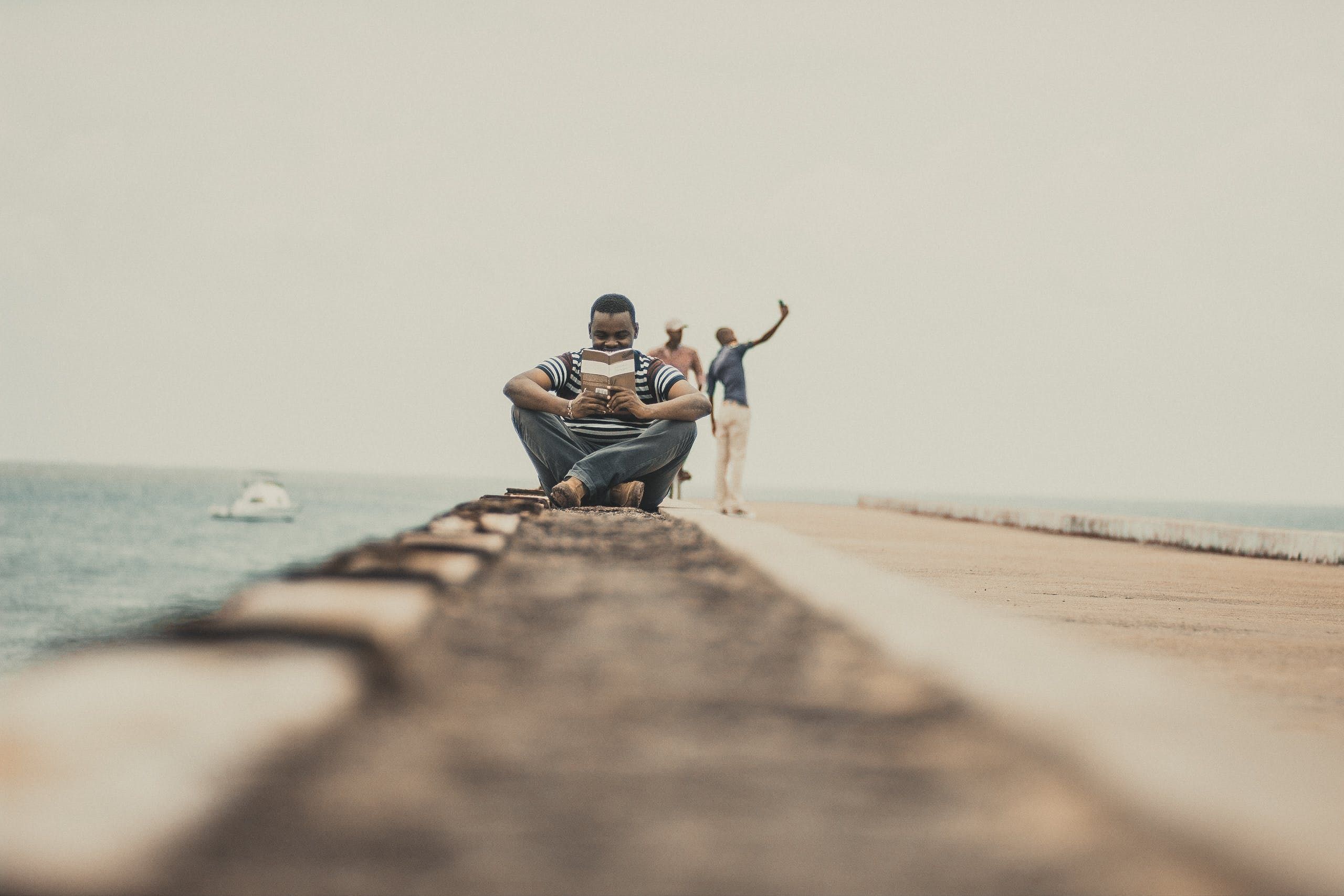 Young man sitting on a seaside promenade, engrossed in reading a book with people in the background taking selfies.