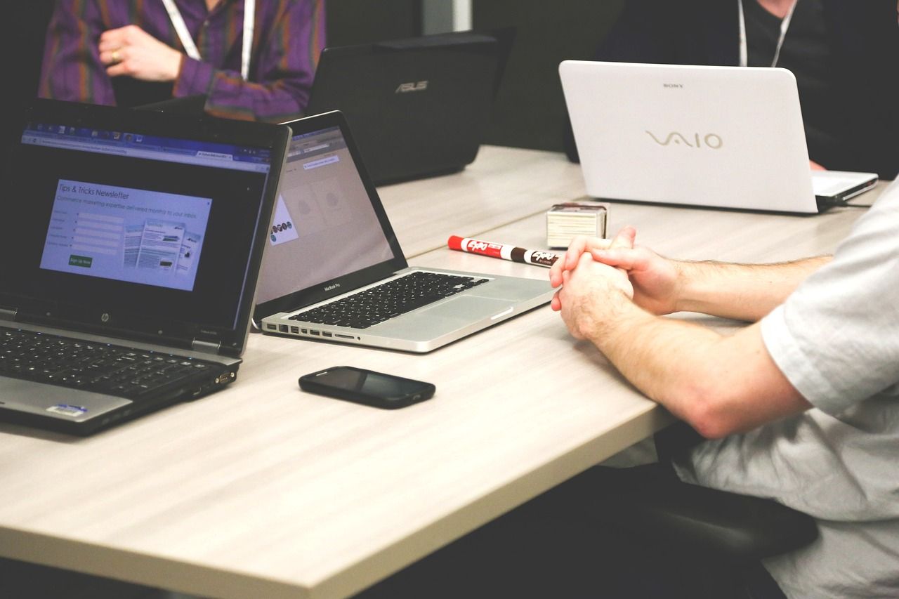 Main Image: Laptops on a desk amidst a meeting at a tech department