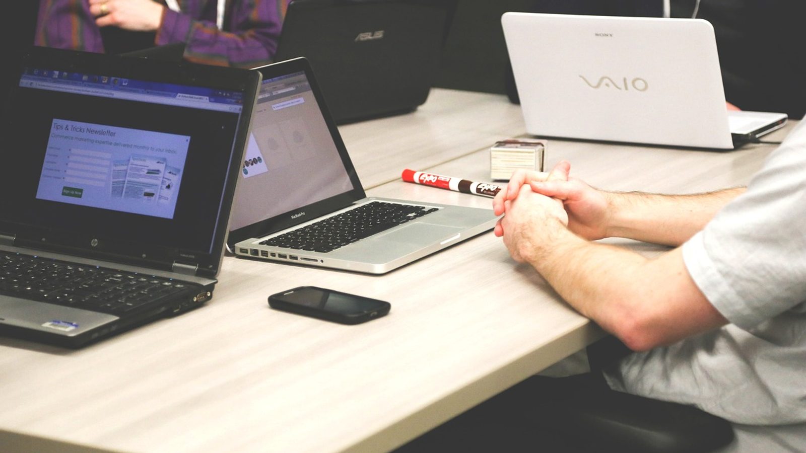 Main Image: Laptops on a desk amidst a meeting at a tech department