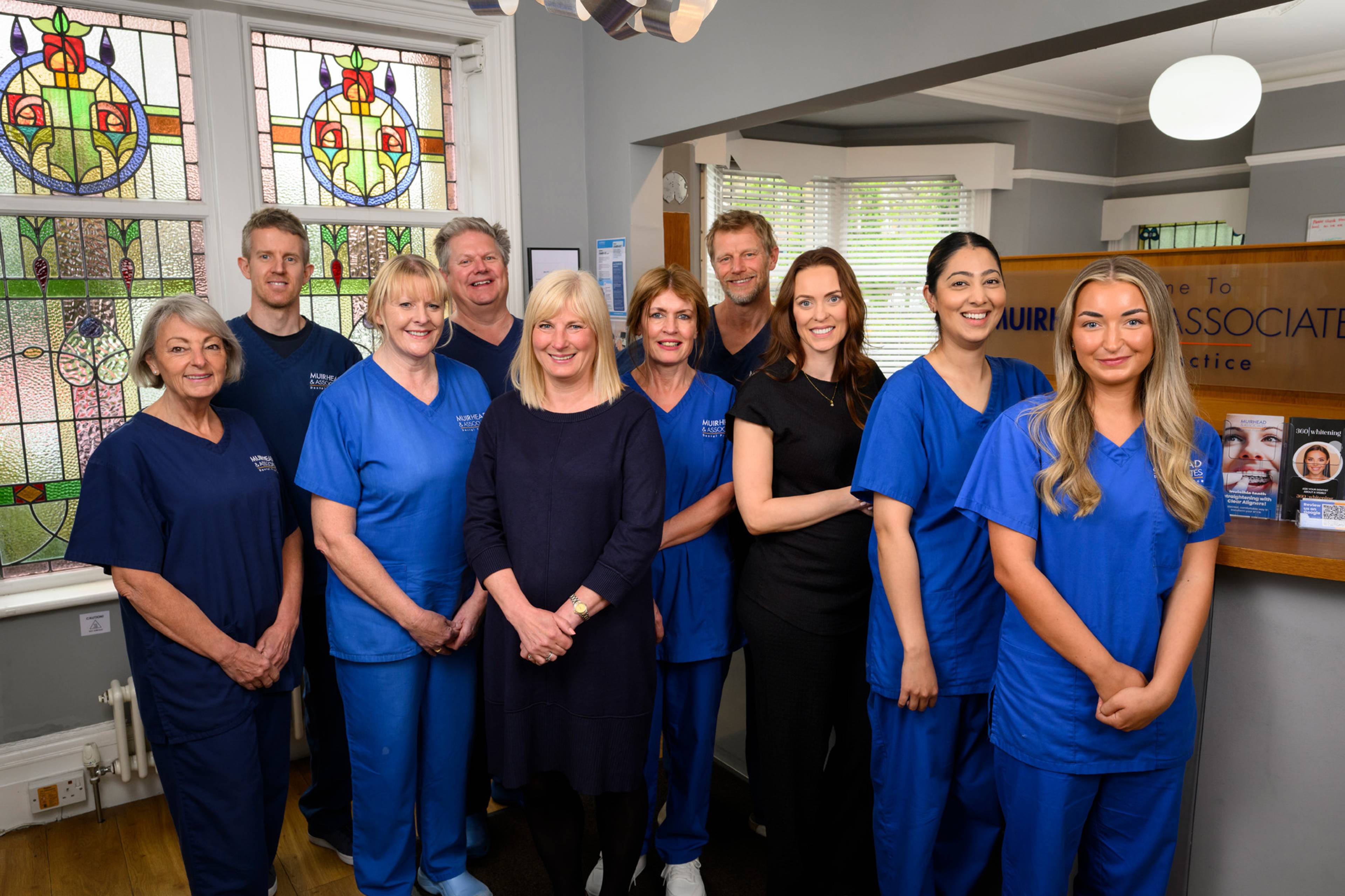 Members of the Muirhead team stand together at the Muirhead & Associates reception area smiling in blue scrubs