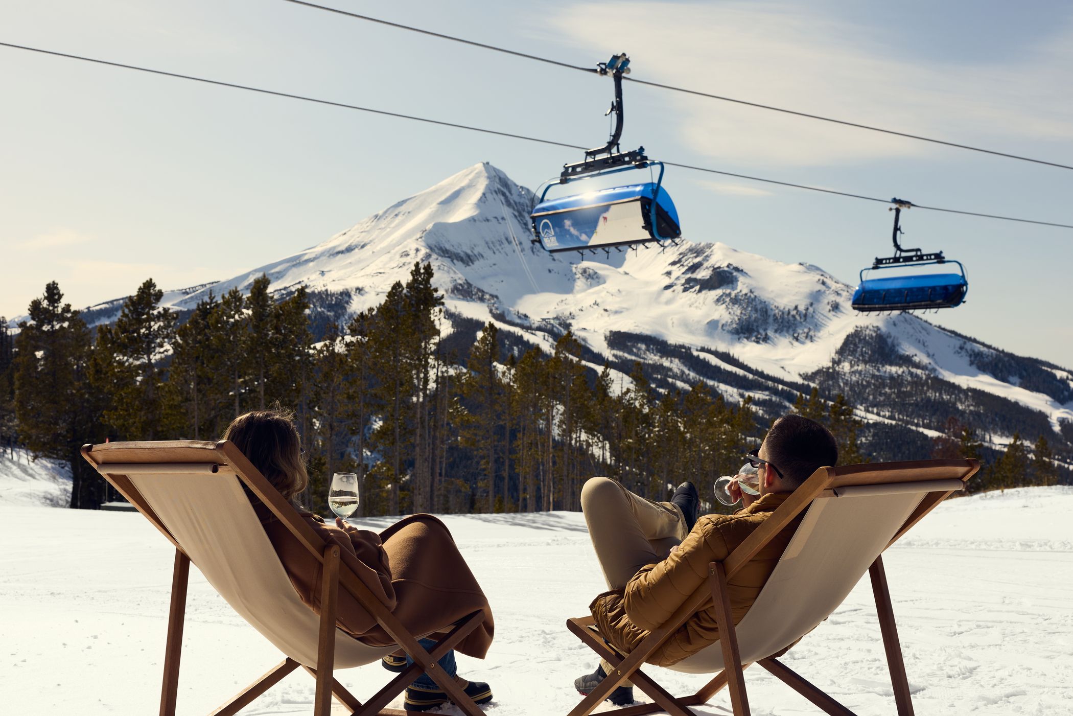People sitting in lounge chairs on the snow with a chairlift in the background