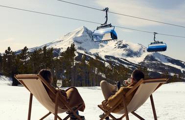 People sitting in lounge chairs on the snow with a chairlift in the background