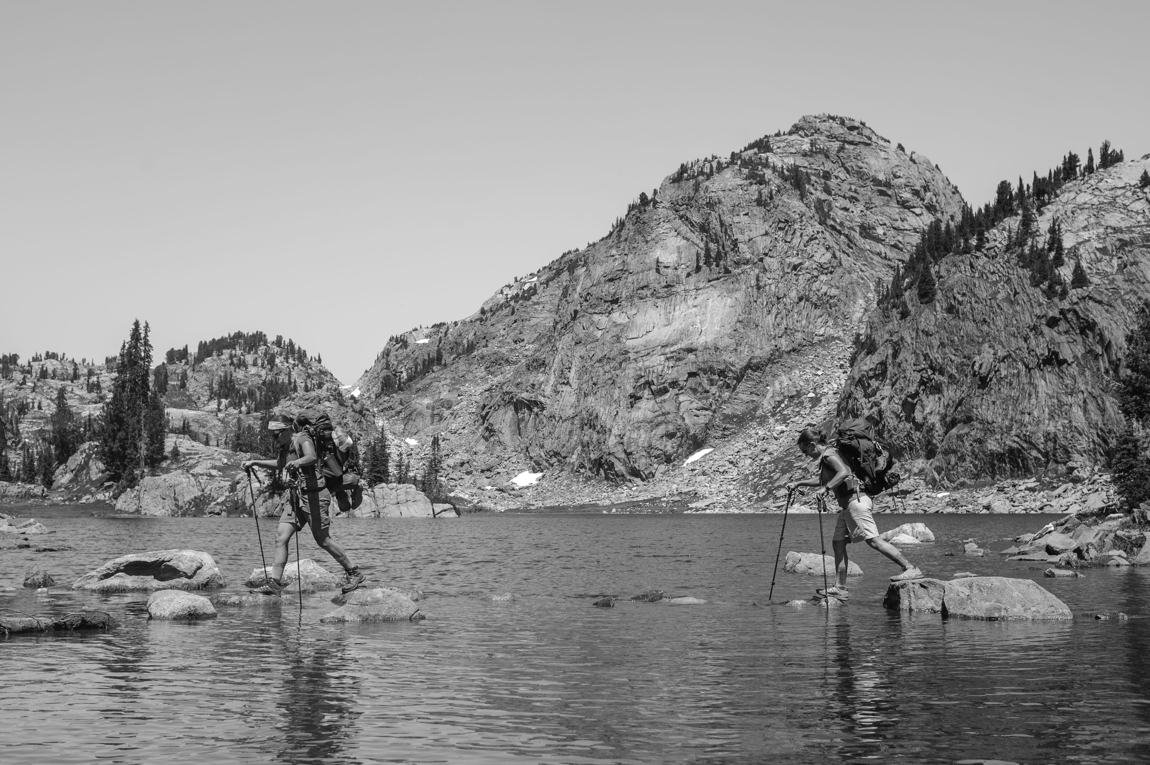 Two women stepping on stones across a lake with backpacking gear