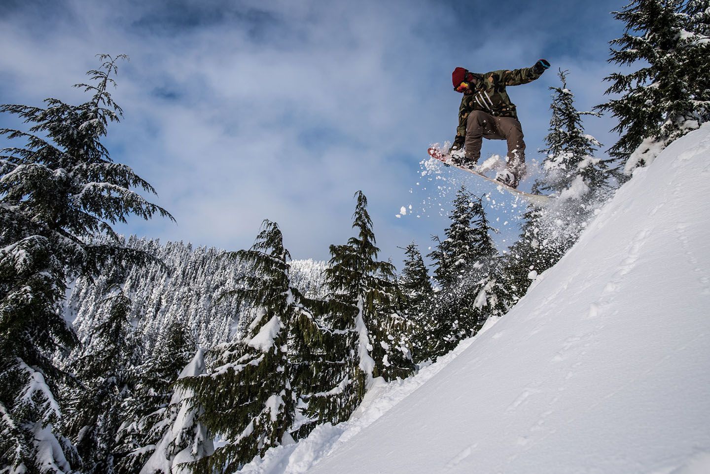 A snowboarder wearing a red beanie, camouflage jacket, and brown pants performs a jump, kicking up snow against a backdrop of tall, snow-covered evergreen trees and a partly cloudy blue sky.