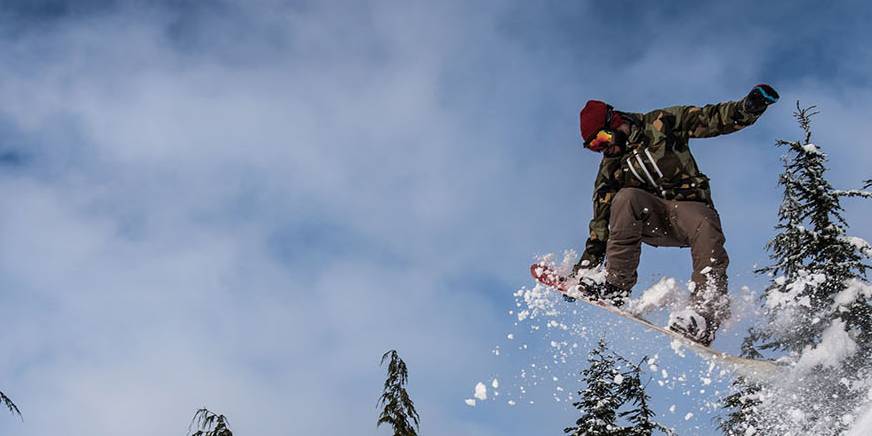 A snowboarder wearing a red beanie, camouflage jacket, and brown pants performs a jump, kicking up snow against a backdrop of tall, snow-covered evergreen trees and a partly cloudy blue sky.