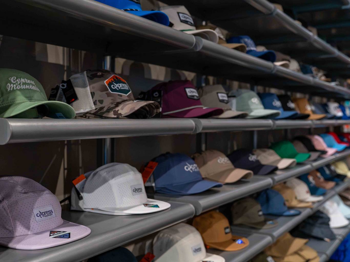 A red hat that says 'Cypress Mountain' hangs on a wall of neck tubes and hats.