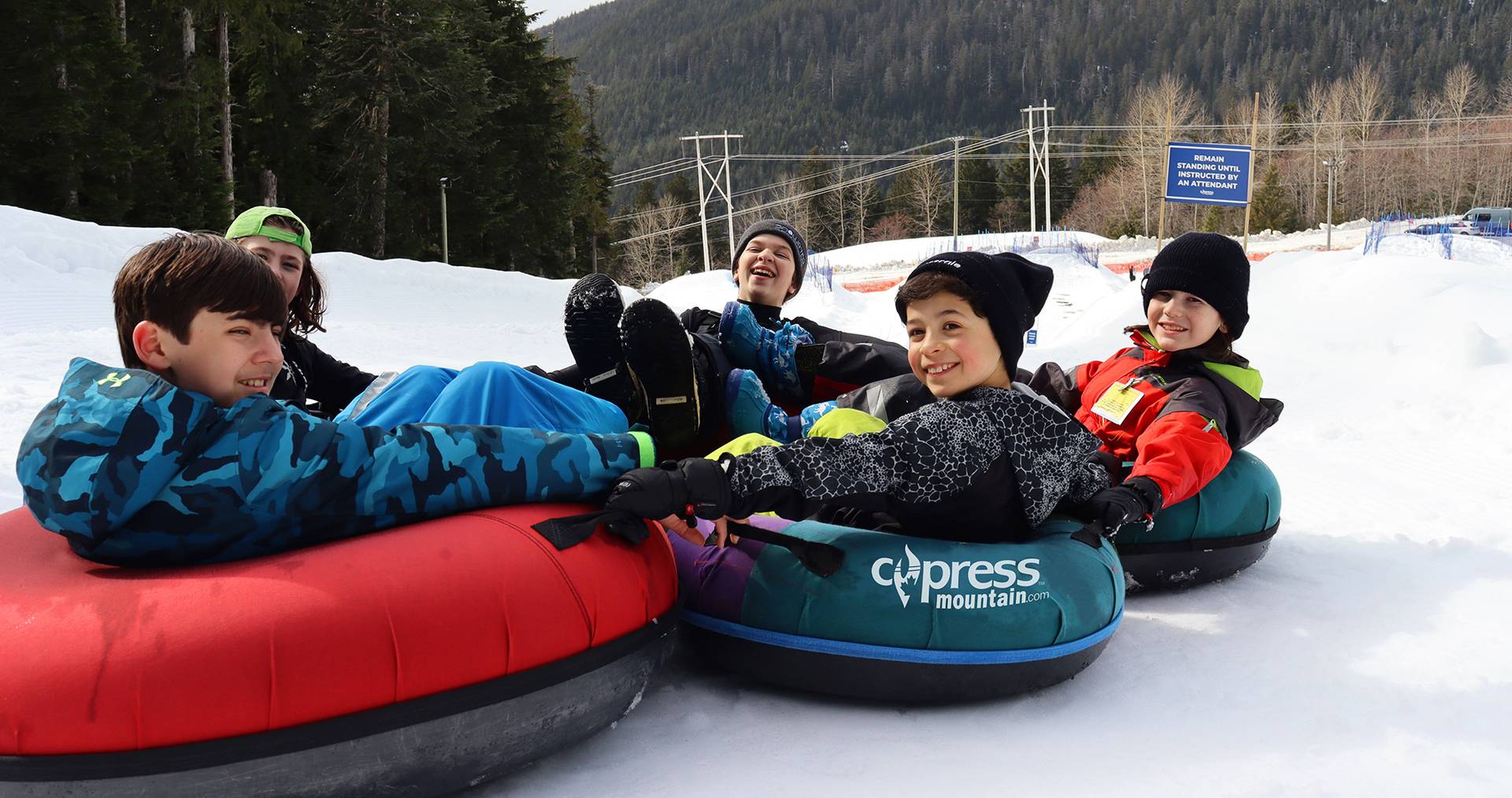 Five young boys wearing multicolored snow suits slide down a tube park while grinning and laughing.