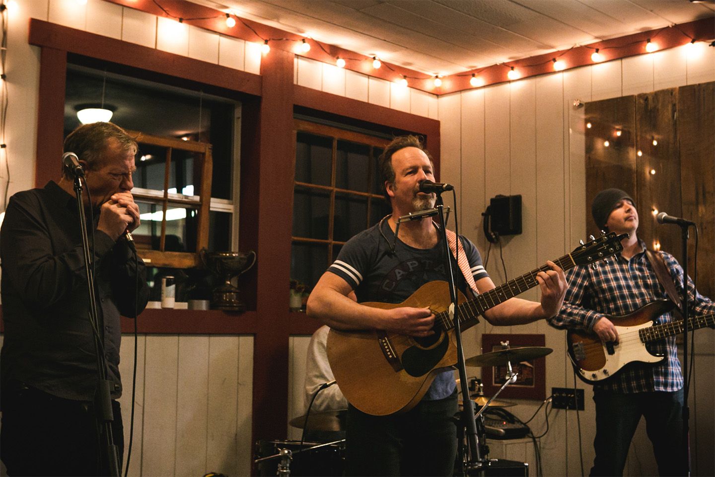 Two men with guitars and one man with a harmonica perform into microphones with string lights behind them.