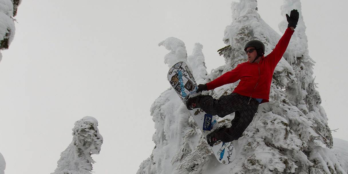 A snowboarder in a red jacket grabs their board while in the air, with a snow-covered tree behind them.