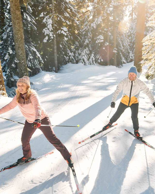 A couple cross country skiing with sun shining in the background through the trees.