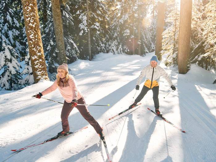 A couple cross country skiing with sun shining in the background through the trees.