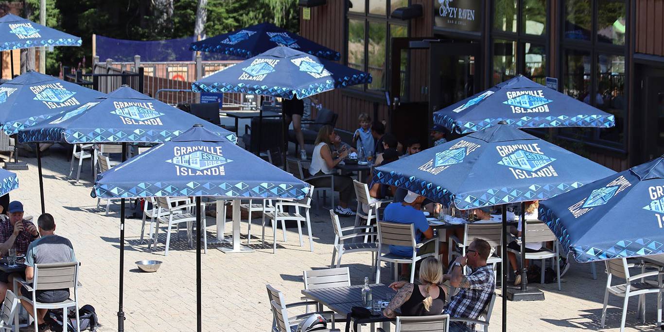 Granville Island umbrellas on a sunny Crazy Raven patio while guests enjoy food and drinks.