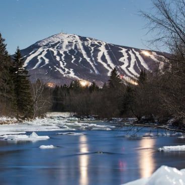 A ski hill with multiple snowy runs, as viewed from the surrounding river. 