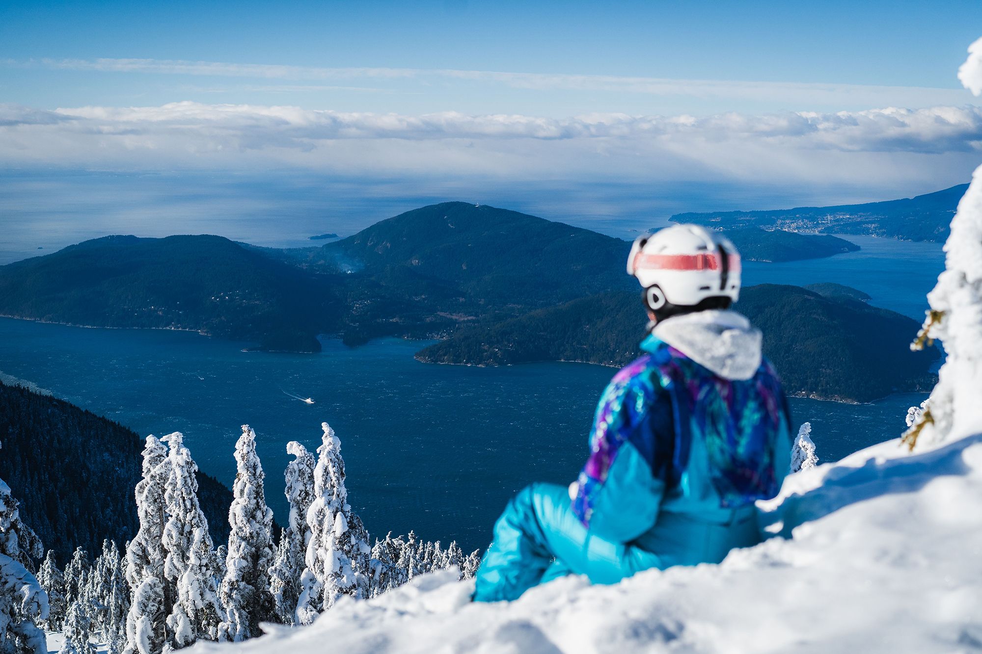 A woman in a blue, retro snowsuit sits on the snow and looks out at the ocean.