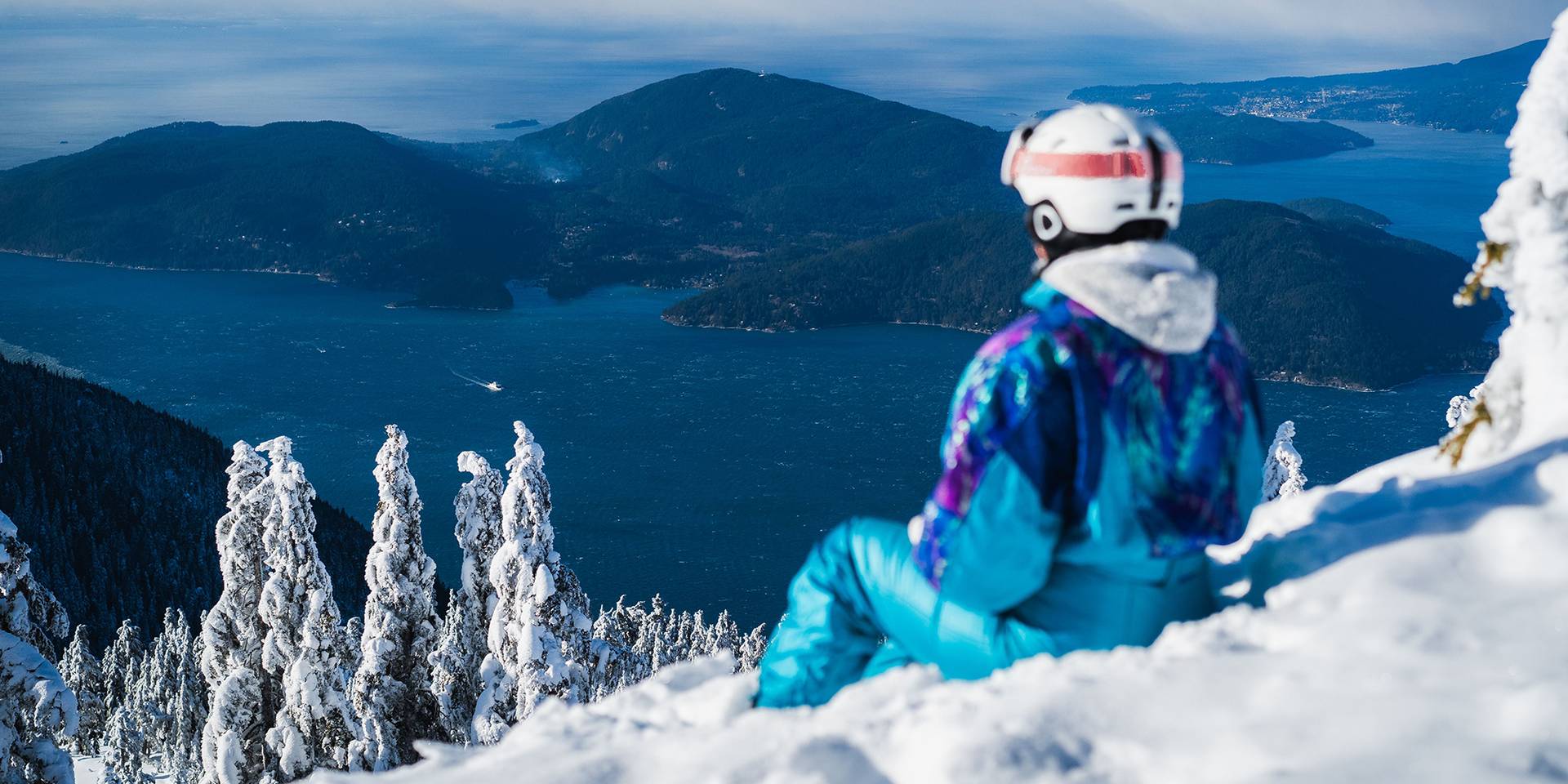 A woman in a blue, retro snowsuit sits on the snow and looks out at the ocean.
