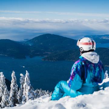A woman in a blue, retro snowsuit sits on the snow and looks out at the ocean.
