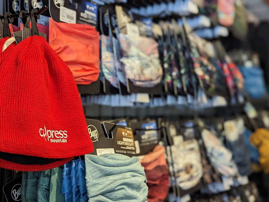 A red hat that says 'Cypress Mountain' hangs on a wall of neck tubes and hats.