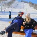 Two skiers seated on a picnic table, drinking coffee, with a ski run in the background.