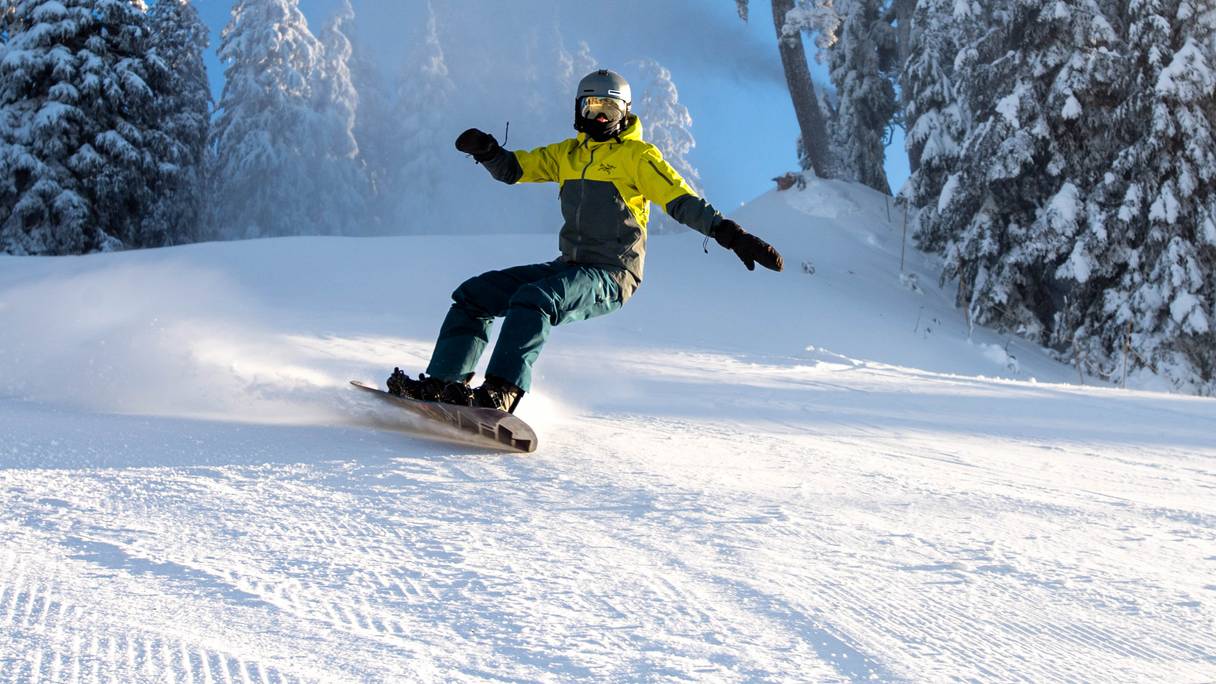 A snowboarder in action, wearing a green jacket on a sunny day.
