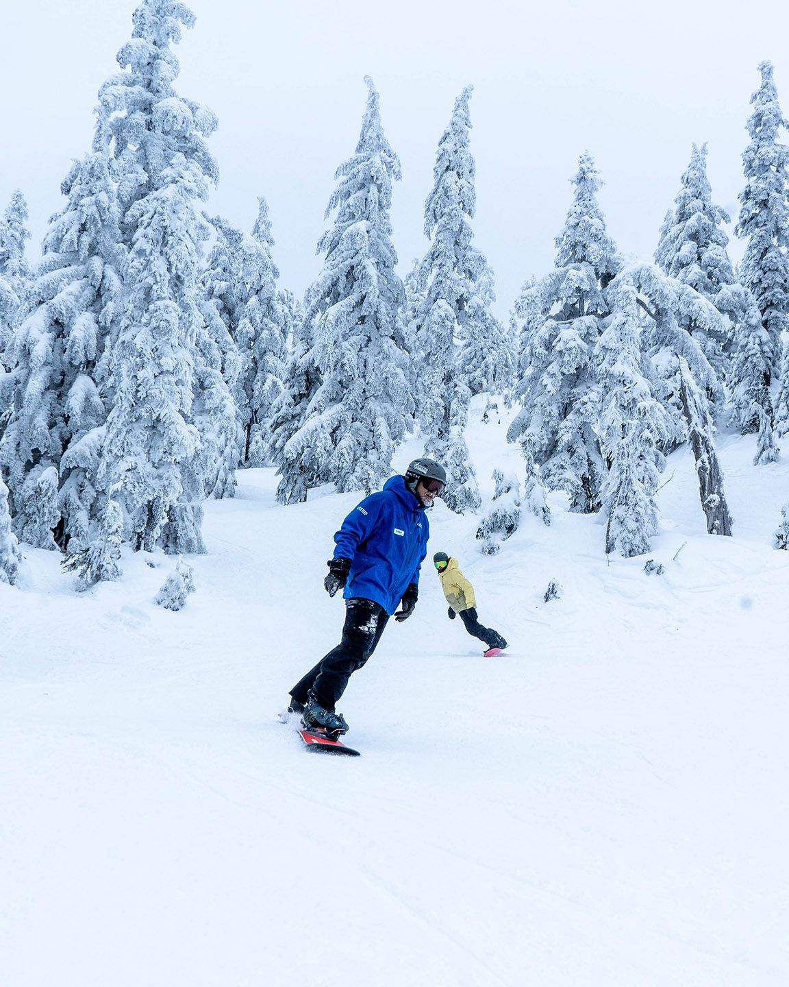 Snowboard instructor in front with student behind with snowy trees in the background.