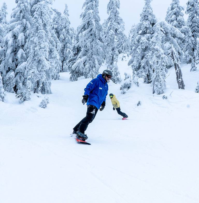 Snowboard instructor in front with student behind with snowy trees in the background.