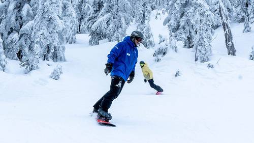 Snowboard instructor in front with student behind with snowy trees in the background.