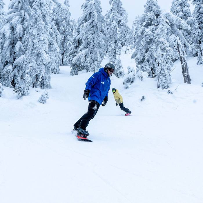 Snowboard instructor in front with student behind with snowy trees in the background.