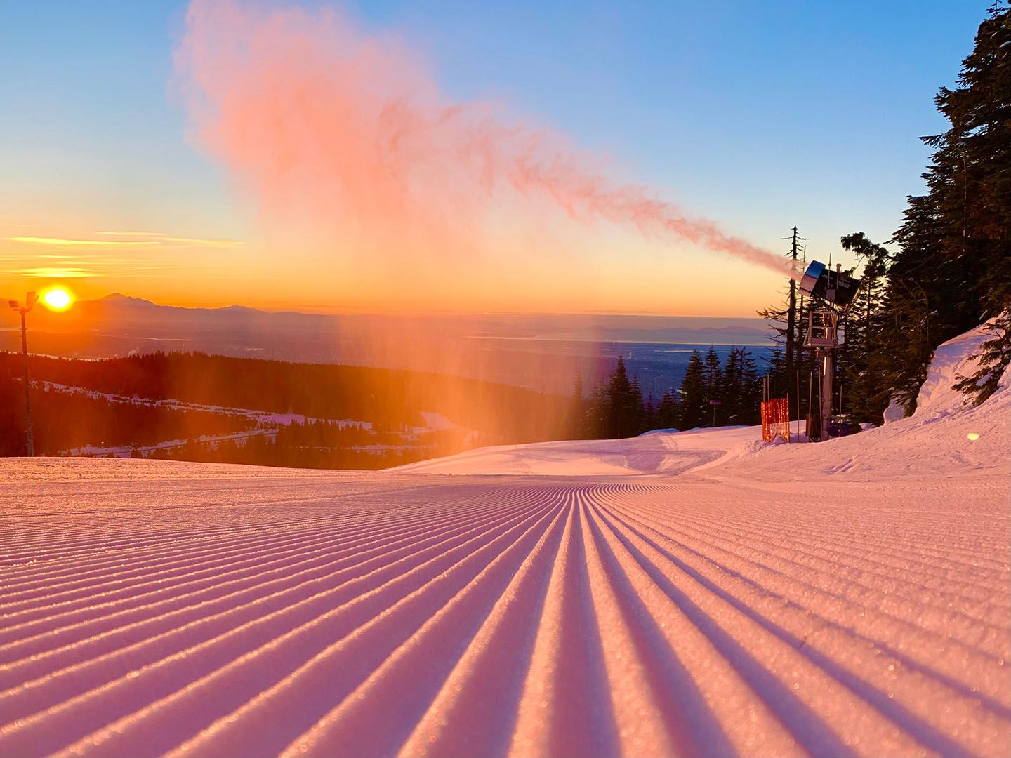 Snow making machines create snow onto a groomed run in front of a sunset in Vancouver.