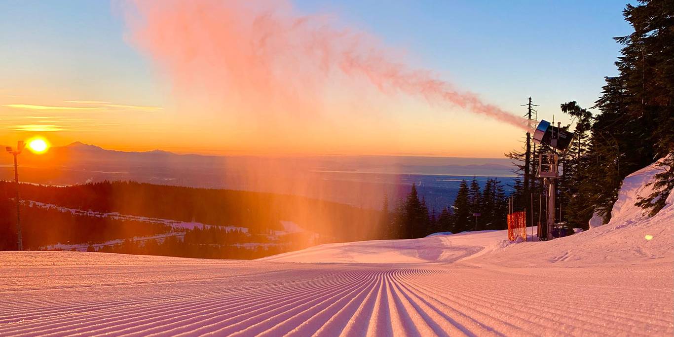 Snow making machines create snow onto a groomed run in front of a sunset in Vancouver.