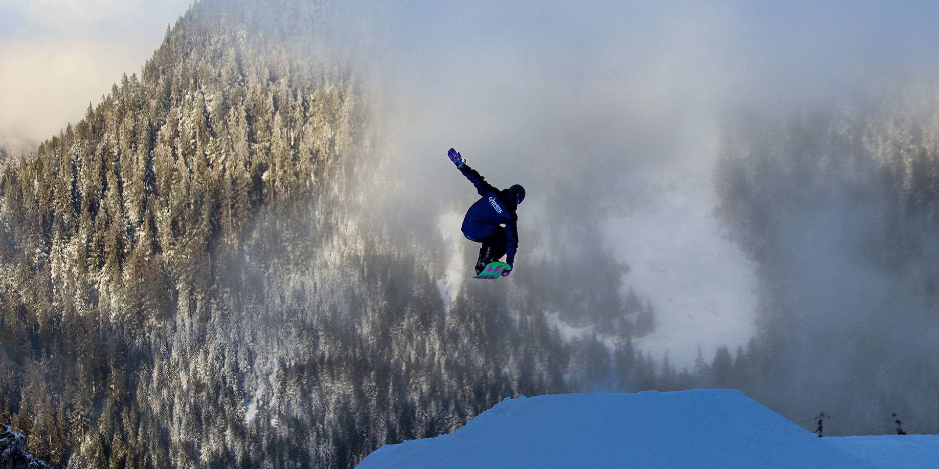 A snowboarder in uniform does a tail grab off a large jump.