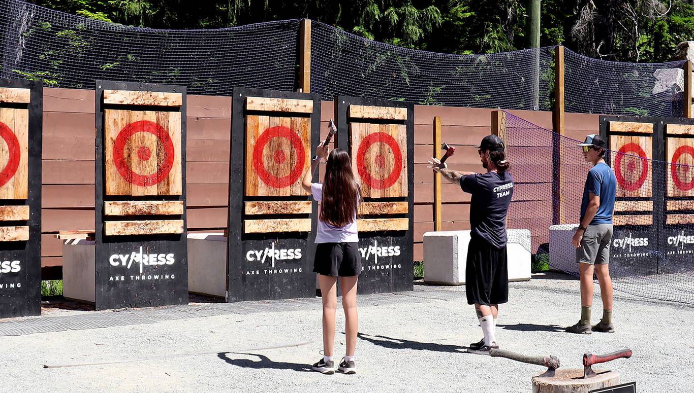Two people axe throw in the mountains while an instructor teaches them.