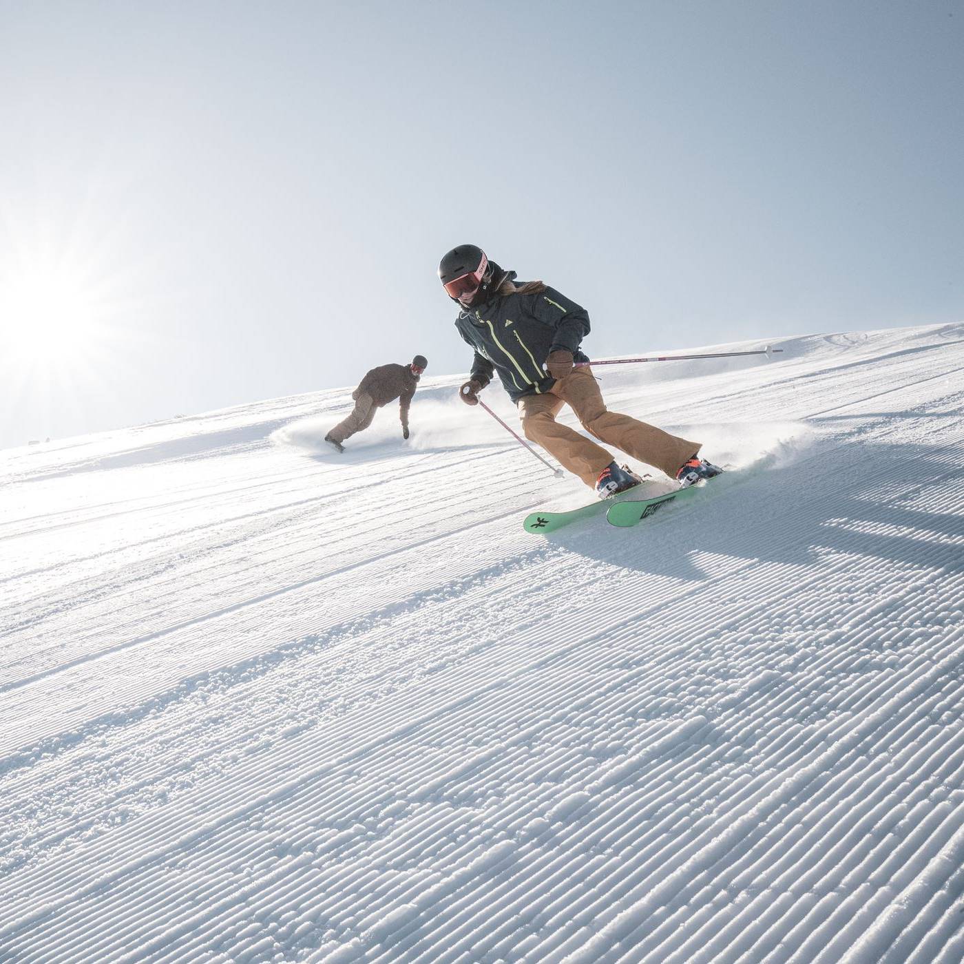 Skier and snowboarder ski down a corduroy groomed run as the sun shines down on them.