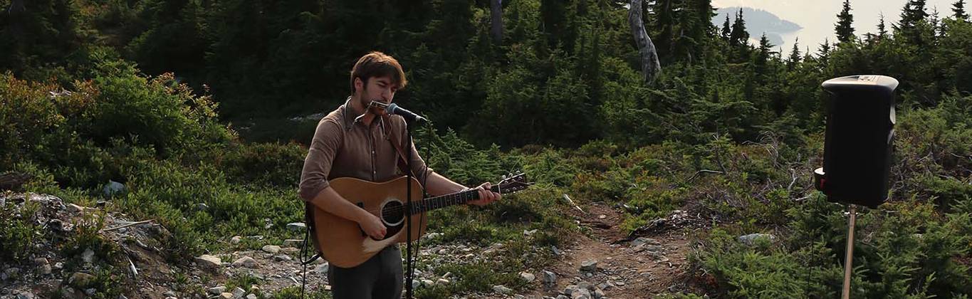 A musician plays an acoustic guitar on top of a mountain as the sunsets behind him.